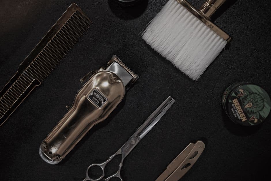 A flat lay of barber tools including a clipper, scissors and comb on a black surface