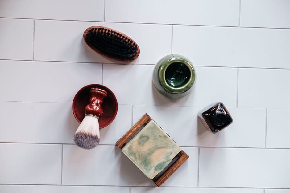 Grooming items laid out on white tile: a brush, a soap in a dish and two jars