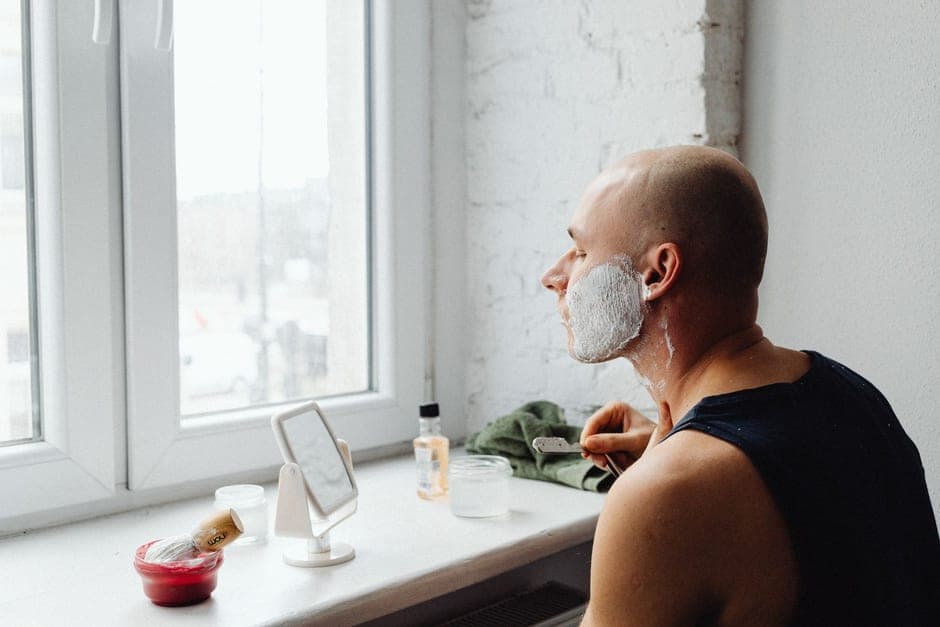A man shaving at a bright window with a mirror and a lather bowl on the sill